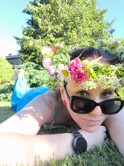 A woman is lying on her stomach in the grass, with a flower crown on her head and sunglasses on. She is wearing a blue dress and a smartwatch. The scene appears to be outdoors, possibly in a garden or park, with a large tree and some bushes in the background. The sun is shining, suggesting it is daytime. The woman is looking directly at the camera with a slight smile. The overall mood of the image is relaxed and cheerful. It's a sunny day in Dzērumi, Latvia.