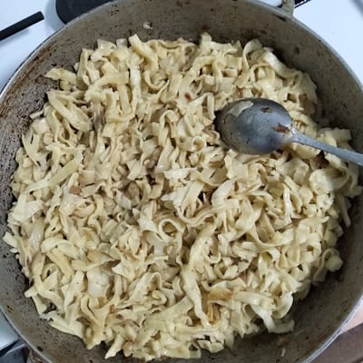 This image shows a close-up view of a pan filled with freshly made pasta. The pasta is light yellow and appears to be a homemade, hand-cut variety with irregular shapes. It is mixed with some browned pieces, likely onions or other aromatics, and glistens with oil or sauce, suggesting it is being cooked or has just been cooked. A metal spoon rests in the pan, partially submerged in the pasta, indicating the food is ready to be served or is in the process of being stirred. The pan itself is well-used, with some dark marks on its interior, suggesting frequent cooking. The background is out of focus but appears to be a kitchen setting, possibly a stovetop, with a white surface and part of a black burner visible. The lighting suggests it is daytime indoors. This scene likely takes place in a home kitchen in Cupcini, Moldova, where homemade pasta is a common dish.