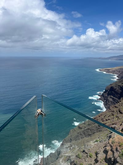 The image captures a scene from the Mirador del Balcón, a viewpoint located in La Aldea de San Nicolás, Spain. The viewpoint features a glass platform that extends out over a cliff, providing a view of the ocean and coastline. The image is taken from the glass platform, looking out towards the ocean. The water is a deep blue color, with white waves crashing against the rocky cliffs. The sky is partly cloudy, with patches of blue sky visible between the clouds. The glass platform is visible in the foreground, with metal supports holding it in place. There are no people visible in the image. The cliff edge is rocky and covered in vegetation.