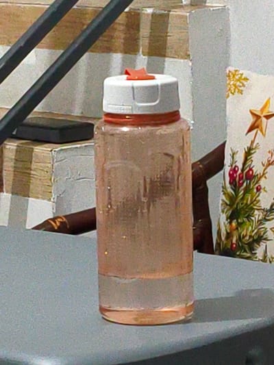 A pink, translucent water bottle with a white and orange lid sits on a gray surface. The bottle is approximately two-thirds full of clear liquid. Behind the bottle, a stack of books with striped covers is visible, topped by a dark rectangular object. To the right of the bottle, a Christmas-themed fabric with a green tree, red berries, and a gold star is propped up. The background consists of a textured, light-colored wall. The scene appears to be indoors, possibly in a residential setting. There is no indication that this scene is located in the city of Cavite City, Philippines.