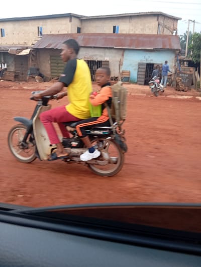 In Imilike, Nigeria, a young Black male wearing a yellow t-shirt and red trousers is riding a motorcycle. A younger Black male, wearing an orange shirt and black pants with an orange stripe, is riding as a passenger behind him. The passenger is carrying a green basket on his lap. They are riding on a dirt road in front of a building with a corrugated metal roof. Another Black male, wearing a blue shirt and jeans, is walking near a motorcycle in the background. The sky is overcast.