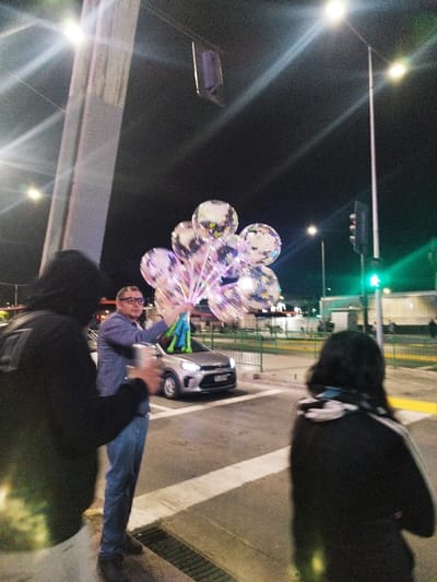 The image depicts an urban street scene at night in Maipú, Chile. Three individuals are visible amidst artificial lighting.

In the center, a male individual, appearing middle-aged, wears eyeglasses, a light blue collared shirt, and jeans. He holds a cluster of approximately eight clear, spherical balloons, each internally illuminated with colorful LEDs. He stands on what appears to be a sidewalk or street median, facing slightly left.

To the left, a person, largely obscured from the chest up, wears a dark hooded jacket and has their back mostly to the viewer. Their left arm is slightly raised, holding an object that is blurred.

To the right, another person, also largely obscured from behind, wears a dark jacket and has dark hair. This individual is positioned as if walking or standing at a pedestrian crossing, facing away from the viewer.

Between the central male figure and the right-side figure, a silver or light grey compact car is stopped on the asphalt road, positioned at what appears to be a pedestrian crossing marked by white lines.

The background features multiple streetlights emitting bright flares, a tall concrete support structure on the far left, and a vertical pole with a traffic light showing a green pedestrian signal on the far right. Distant background elements include indistinct buildings and green metal barriers. The overall scene is illuminated by various artificial light sources, creating strong highlights and shadows. The image exhibits visible noise consistent with low-light photography.
