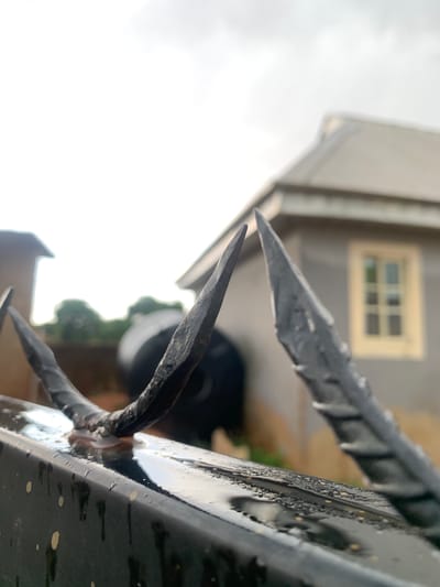 The image shows a close-up of a black metal fence or gate with sharp, pointed spikes on top. The metal is wet, possibly from rain. In the background, a blurred building with a window is visible. Based on the city provided, the image is most likely located in Farin Yashe, Nigeria. There are no people visible in the image. The focus is on the fence as a security measure. The environment appears to be residential.