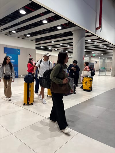 In an indoor, modern airport terminal in Callao, Peru, a group of travelers are seen walking through a wide, well-lit corridor. The floor is tiled with large, light-colored squares. Overhead, a suspended ceiling with exposed dark beams and red piping is visible, interspersed with numerous white, circular lights.
In the foreground and midground, several individuals are navigating the space with luggage. On the left, a woman with dark, long hair wears a grey hooded sweatshirt and beige pants, carrying a small black crossbody bag. Beside her, a yellow wheeled suitcase stands upright. Further back, a man in a white t-shirt and a white baseball cap is seen from the chest up, holding a cell phone. He is accompanied by a person in a red jacket holding a phone and a person in a blue vest. Behind them, a large black backpack is placed on a yellow wheeled suitcase.
In the mid-right of the image, a woman with dark hair is walking away from the camera, wearing a dark olive-green long-sleeved shirt and black pants, carrying a brown shoulder bag. To her right, a yellow duffel bag with "THE NORTH FACE" branding is visible. Near a large concrete pillar in the mid-right, two individuals are interacting, one wearing a dark blue uniform and writing on a clipboard, and another in a striped shirt. Beyond them, glass doors and more terminal architecture are visible. Another yellow suitcase is partially visible on the far right. The overall atmosphere suggests transit and movement within an airport environment.
