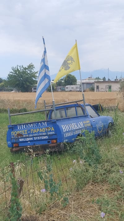 A faded blue pickup truck, likely abandoned, sits partially submerged in tall, dry grass and weeds, with a rusty wire fence in the foreground. The truck's bed is adorned with two flags: a Greek flag on the left, and a yellow flag with a black eagle on the right, possibly symbolizing the Byzantine Empire. The truck's side displays text in Greek, "BIOMKAM ΠΟΡΤΕΣ - ΠΑΡΑΘΥΡΑ" (BIOMKAM DOORS - WINDOWS) and "MALUMIN SYSTEMS", along with phone numbers. The background reveals a rural landscape with dry fields, trees, and a few distant buildings under a cloudy sky. The scene is set in Νέα Κερασιά, Greece, suggesting a location possibly on the outskirts or in a less maintained area of the town.
