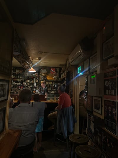 The image depicts the interior of Matt Molloy's Yard Bar in Westport, Ireland, a traditional Irish pub. The dimly lit atmosphere is characteristic of such establishments, with warm lighting from a pendant lamp above the bar casting shadows. The bar itself is well-stocked with numerous bottles of alcohol, neatly arranged on shelves behind the bar. Several patrons are visible, some standing at the bar and others seated. A bartender, a young man with reddish-blond hair, is serving customers. The walls are adorned with a variety of framed pictures and posters, contributing to the pub's character. An air conditioning unit is mounted high on the wall, and an illuminated green emergency exit sign is visible. The overall scene conveys a lively yet intimate pub environment.