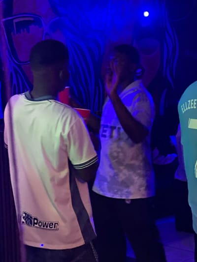 The image depicts an indoor scene with several individuals, predominantly young Black males, socializing in what appears to be a nightclub or party setting. The environment is characterized by prominent blue lighting and a mural or painted backdrop featuring stylized facial features and abstract designs.
In the foreground, a young man wearing a white t-shirt with a dark stripe on the sleeve and a logo reading "Power" is partially visible from the back. To his right, another young man in a light-colored, possibly tie-dye shirt, is facing towards the right and gesturing with his hand near his face. His shirt has text on it that is partially obscured. To the far right, a portion of a third individual's light blue t-shirt is visible, displaying the text "ELLZEE".
The people appear to be engaged in conversation or enjoying the ambiance of the location. Red cups are visible, suggesting the presence of beverages. The overall impression is one of a lively social gathering. The location within the city of Durumi, Nigeria is not explicitly indicated by any visual cues in the image itself.