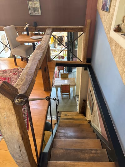 This interior shot depicts an elevated perspective of a room with a blend of rustic and modern elements. A heavy, exposed wooden beam framework is prominent in the foreground, with an attached small round table set for one with a wine bottle and glass. The view extends towards a staircase constructed of wood treads. In the background, a glimpse of an outdoor scene, potentially a street or shop, is visible. A red patterned rug adorns the floor, and the surrounding walls feature a mix of materials, including wood paneling and painted surfaces. The location is likely an interior space within the city of Bartula, Montenegro, identifiable by the distinctive architectural style and decor.