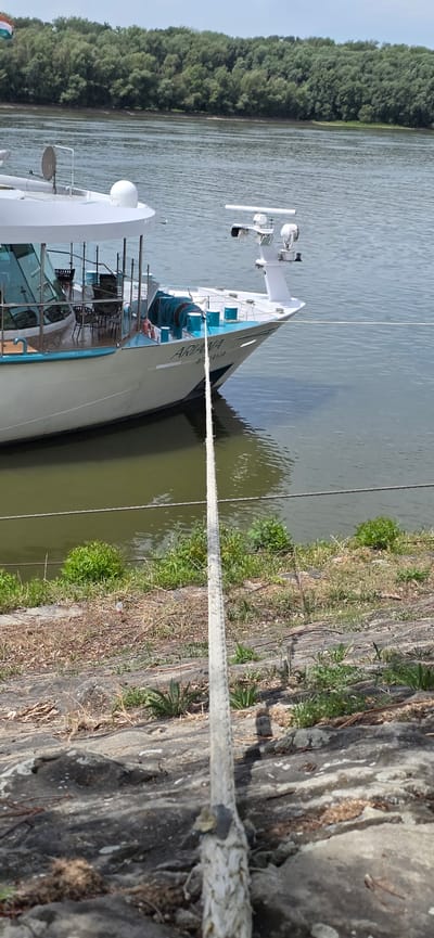 The image captures a moored white ship named "ARIANA" on a body of water, likely a river, in Foktő, Hungary. The ship is white with turquoise trim and has a white superstructure. Various nautical equipment, including radar, antennas, and lights, are visible on the ship's deck. Two thick ropes tether the ship to the shore. The foreground shows a rocky, overgrown embankment with sparse vegetation. In the background, a dense line of green trees borders the water, forming a natural landscape. The sky is mostly clear with some faint clouds. There are no people visible in the frame, and the scene appears static, suggesting the ship is docked.