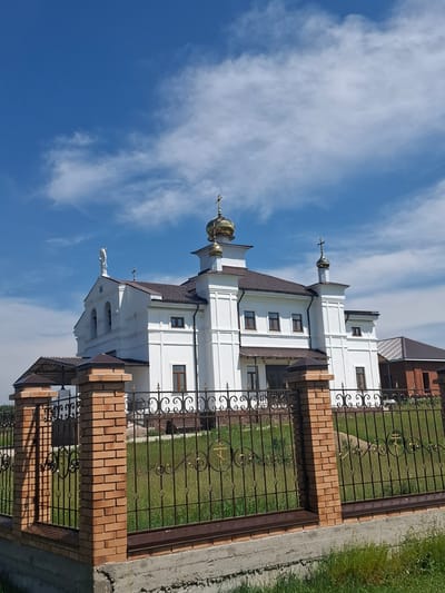 The image shows a white Orthodox church with a golden dome, set against a blue sky with scattered clouds. A brick and metal fence encloses the property. The church features multiple windows and crosses on its roof. To the right of the church, a red brick building with a dark roof is partially visible. The ground within the fence is covered in green grass. The foreground shows a concrete foundation and more green grass. The location is identified as Bayanday, Russia.