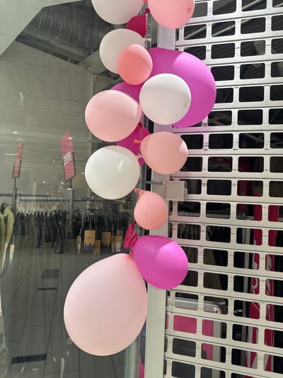 This image shows a collection of pink and white balloons clustered together, suspended in front of a silver grate-like structure. The balloons vary in size and shade of pink, with one prominent white balloon. The grate structure appears to be a decorative element, possibly part of a store display or architectural feature, with dark rectangular openings.

Reflected in the glass surface below the balloons, we can see rows of clothing on hangers, suggesting the setting is a retail environment. There are also visible price tags and signage. One sign reflects a "399" with a reverse image of the numbers. The overall lighting suggests an indoor environment, possibly a store during operating hours. There are no people visible in the image, but the presence of balloons and the retail setting imply a celebratory or promotional event, or simply store decoration. The context of Slitu, Norway, is provided but not visually evident within the image itself.