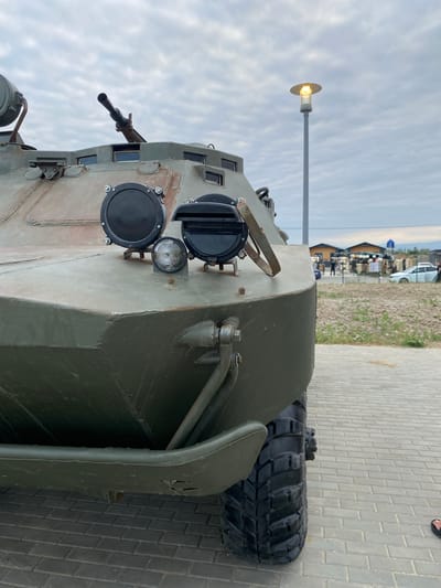 The image shows the front of a military vehicle, possibly an armored personnel carrier, parked outdoors on a paved surface. The vehicle is painted olive drab and features a prominent machine gun mounted on top. Several lights and observation ports are visible on the front. In the background, there is a lamppost, some buildings, and people milling around, suggesting a public area or event. The sky is overcast. The ground is paved with grey bricks. The vehicle is positioned at a slight angle to the camera, with its front left tire visible.