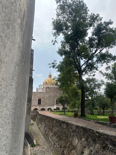 The image captures an outdoor scene in Tepotzotlán, Mexico, featuring historical architecture and natural elements under a bright, partly cloudy sky. The perspective is low, looking over a textured, grey concrete or stone ledge that occupies the bottom portion of the frame.
In the midground, a substantial stone wall with a brick top runs horizontally, separating the foreground from a lush green area. Beyond this wall, a paved pathway is visible, leading to a garden or courtyard with several trees and grassy patches.
The dominant feature in the left half of the image is a large, leafy tree with dark green foliage, its branches extending towards the sky. Through the trees and above the midground wall, a prominent stone building with a striking golden dome and a bell tower is visible. The building's walls appear to be constructed from dark, rough-hewn stone, suggesting historical significance. The golden dome, with a cross at its apex, gleams brightly, indicating the building is likely a church or a cathedral, possibly part of the Museo Nacional del Virreinato. Arched openings are discernible on the side of the building.
The scene appears peaceful and devoid of any human activity. The time of day is daytime, with soft, ambient lighting suggesting either late morning or early afternoon. No visible text is present in the image.