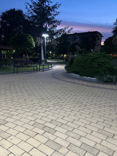 This image depicts a public park or pedestrian area in Dzhebel, Bulgaria, during twilight or early night. The foreground is dominated by a light-colored paved pathway composed of rectangular interlocking blocks, extending from the bottom of the frame into the mid-ground. The pathway curves to the right, where a section is bordered by darker, reddish-brown blocks. To the left of the main path, two dark-framed park benches with wooden slats are situated on a strip of grass.
In the mid-ground, a modern street lamp with a flat, circular top emits bright white light, illuminating the immediate area. Beyond it, various trees, including a tall coniferous tree with dark, dense foliage and a lighter, bare trunk visible beneath its canopy, are silhouetted against the sky. A smaller, rounded, brightly lit green tree stands to the left of the coniferous tree. Another street lamp is visible further in the distance to the right, along with a partial view of a red circular sign.
The background reveals a multi-story building, partially obscured by trees and foliage, appearing somewhat weathered, with antenna-like structures on its roofline. The sky above is a gradient of deep blue, transitioning to a strip of pink and purple hues along the horizon. Two faint celestial objects (likely stars or planets) are visible in the darker blue portion of the sky. Dense green shrubbery and smaller plants line the pathways, with a cluster of bushes in the mid-right featuring white decorative stones at its base. A dark, gazebo-like structure is partially visible on the far left. No individuals are present in the scene.