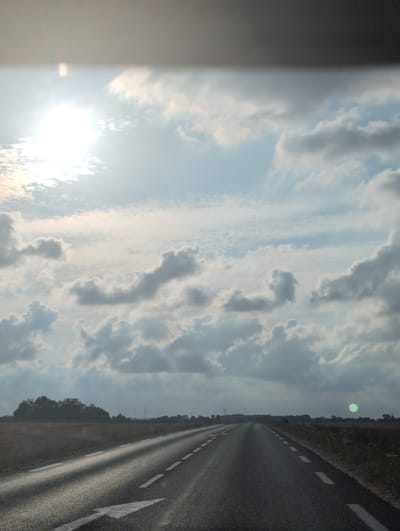 The image depicts a road perspective in Bracquetuit, France, on a cloudy day. The road, asphalt, stretches into the distance, marked with white dashed lines and a solid white arrow indicating a lane. Fields flank the road, with a line of trees in the mid-ground. In the background, wind turbines are visible. The sky is filled with dramatic clouds, with the sun peeking through on the left, creating lens flare. A faint green orb is present in the sky on the right side of the image. There are no people visible in the frame.