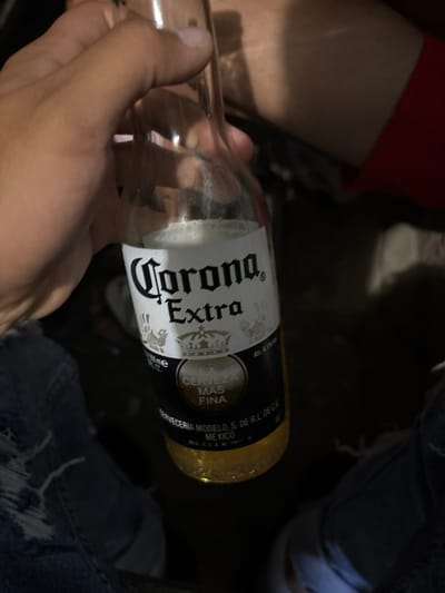A person's hand is holding a clear glass bottle of Corona Extra beer. The bottle is mostly full of yellow beer and has a white label with "Corona Extra" written in black text. The label also shows a crown and other ornate designs. The background is dark and appears to be an indoor setting, possibly a dimly lit bar or restaurant. The image is taken from a close-up perspective, focusing on the bottle and the hand holding it. There is no discernible information about the location in the city of Gorno Aleksandrovo, Bulgaria.