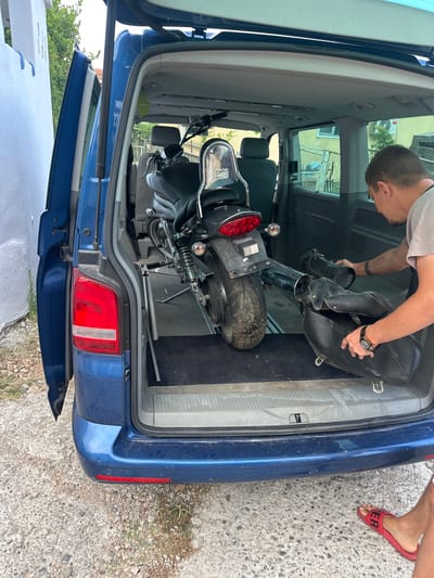 A man is standing at the rear of a blue Volkswagen Transporter van with its rear hatch and side door open, in the city of Đinđinovići, Montenegro. The van's interior is equipped for transporting a motorcycle. A motorcycle, possibly an electric scooter or a small motorcycle, is loaded into the van, positioned lengthwise in the cargo area. The man is holding black motorcycle saddlebags, one in each hand, and appears to be placing them into the van. The background shows a white building on the left and a fence and trees on the right, suggesting a residential or industrial area. The ground is covered with gravel and concrete. The man is wearing a gray t-shirt and dark shorts. His feet are clad in red slide sandals. The lighting indicates it is daytime, possibly late morning or afternoon.