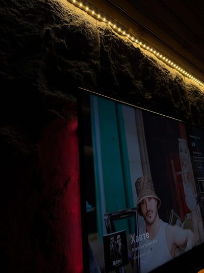 The image shows a wall covered with rough plaster, divided into two sections by different lighting. The upper left portion is lit by a strip of warm white LED lights, illuminating the textured surface. The lower right area is illuminated with a red light source.
Mounted on the wall is a flat-screen television, displaying the song "Хавте" by Tsvetelina Yaneva and Azis, a Bulgarian song. On the screen is a male figure wearing a patterned bucket hat and a white t-shirt. There is also an album cover with the title "Хавте" at the bottom right of the screen.
The overall scene suggests a room interior with custom lighting. The city of Lilkovo, Bulgaria, is not clearly identified in the scene.