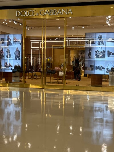 A Dolce & Gabbana store front is reflected in the shiny floor. The facade has gold trim, with "DOLCE & GABBANA" vertically displayed on the left. Inside, clothing and accessories are visible. Two video displays on the left showcase shoes and handbags. Customers in patterned dresses are seen inside.