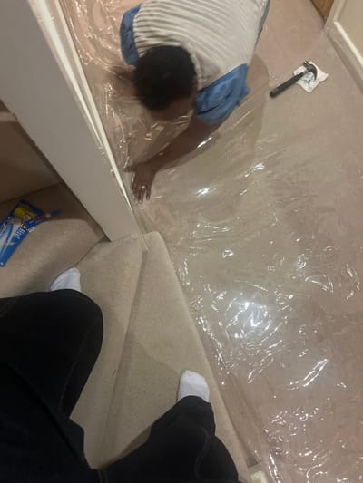 This image captures an indoor scene within a residence in Stevenage, United Kingdom, showing two individuals involved in a domestic activity.
One person, with dark, short hair, is depicted on their hands and knees on a floor covered with clear plastic sheeting. This individual wears a horizontally striped grey/white short-sleeved top over a blue undershirt or sleeve, and a ring is visible on their left hand. Their arms are extended forward, with hands near the base of a white vertical banister or newel post, suggesting they are working with the plastic sheeting or the underlying floor.
To the right of this individual, a black-handled claw hammer rests on a crumpled piece of white material, possibly a cloth or paper. The floor beneath the plastic sheeting appears to be a light-colored surface, possibly carpet or wood.
In the foreground, the lower half of a second person is visible, encompassing their legs clad in dark trousers and feet in white socks. This person appears to be positioned on carpeted stairs, which are a light beige color, looking down towards the scene.
To the left of the white banister, on the carpeted floor next to a wall, a blue plastic bottle with white lettering, possibly a cleaning product, is present. The overall environment suggests a renovation, cleaning, or protection task in progress within a residential hallway or landing area.