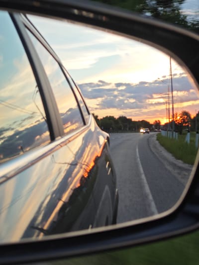 The image captures a vibrant sunset reflected in the side-view mirror of a car. The primary subject is the view through the mirror, which shows a road curving to the right. In the distance, another car's headlights are visible, appearing to approach. The sky is filled with a beautiful sunset, displaying warm orange, pink, and yellow hues along the horizon, contrasted with some blue and gray clouds higher up. The dramatic colors of the sky are also reflected on the sleek, metallic body and window of the car from which the photo is taken, creating a sense of dynamic light and movement.

Along the right side of the road in the reflection, several utility poles or streetlights stand, accompanied by lush green grass and trees lining the roadside. The overall setting suggests an evening drive through a rural or suburban area. The frame of the side-view mirror is dark, framing the captivating scene within. The activity depicted is traveling by car during dusk. There is no visible text in the image. The location is Fedino, Russia.