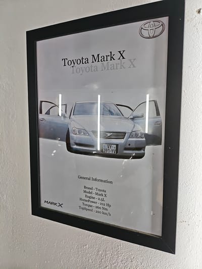 A framed black and white poster is displayed on a textured white wall. The poster features a silver Toyota Mark X sedan with its driver's side door open. The car is positioned centrally, with its front end prominent. Above the car, "Toyota Mark X" is printed twice, once in black and once in a lighter gray, slightly blurred. The Toyota logo is visible in the upper right corner of the poster. Below the car, text details the car's specifications: "General Information," "Brand - Toyota," "Model - Mark X," "Engine - 2.5L," "HorsePower - 212 Hp," "Torque - 260 Nm," and "TopSpeed - 220 km/h." The bottom left corner of the poster has a logo that reads "MARK X." The image was taken in Krtsanisi, Georgia.