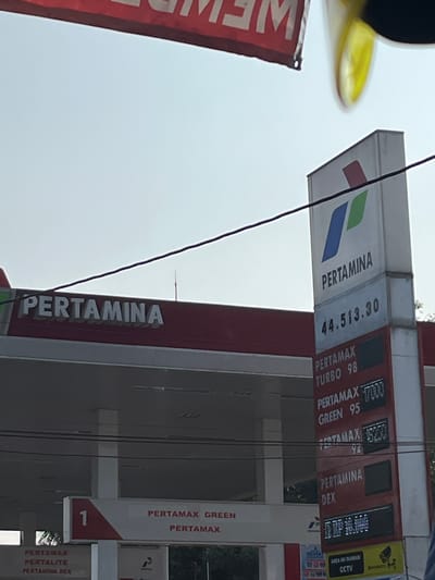 This media file captures a vertical shot of a Pertamina gas station in Boja, Indonesia. The main subjects are the prominent signage and pricing displays of the gas station. Multiple fuel types are advertised, including Pertamax, Pertamax Turbo 98, Pertamax Green 95, and Pertamina Dex. The prices are displayed digitally, with one visible price reading "44.513.30".

The scene appears to be during daylight, with a bright, overcast sky visible in the background. A red banner with white text, partially obscured and unreadable due to its orientation and blurriness, is visible on the left. A yellow sign with the text "AREA INI DI AWASI CCTV" (This area is monitored by CCTV) is present on the right, indicating security measures.

The overall impression is a functional and busy gas station environment. The image is slightly tilted, adding a dynamic perspective to the otherwise static display of information. No people are directly visible, but the presence of a CCTV sign suggests a public space. The concrete and metal structures of the gas station, along with the hanging wires, are characteristic of such infrastructure in Indonesia.