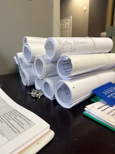 The image shows a stack of rolled-up papers, likely documents, on a dark surface. The top roll is labeled "Monthly Behavior Oversight." Several other rolls have printed information visible, including "Rx # 1006183-0," "Patient Name: WIL," and "Address: 4771." A blue Primacare item and a stack of papers with a green binder are positioned behind the rolls. In the foreground, a partially visible document shows text, including "MODUPE DAWODU" and dates. The environment appears to be an office or workspace. The city of Loganville, United States of America, is not directly evident in the image.