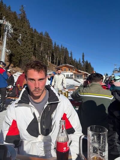 The scene depicts an outdoor area at a ski resort, identified by a building with "HOLLENAU HÜTTE" visible, situated in Schwendau, Austria. The weather is clear and sunny with a bright blue sky.

In the foreground, a male individual with short, curly brown hair and a mustache is seated at a light wooden table, wearing a white, black, and red ski jacket over a grey t-shirt. He looks directly at the camera with a neutral expression. A glass bottle of Coca-Cola with a red label is positioned in front of him on the table. To his right, a large, clear glass beer stein, showing residue of consumed liquid and foam, is also on the table.

To the right of the foreground subject, another male is seated at the same table, with his back to the camera. He wears a dark green ski jacket with two horizontal yellow and white stripes across the upper back.

In the mid-ground, numerous individuals dressed in various ski attire are visible. Some are standing, and others are seated at additional wooden tables. The Hollenau Hütte building, a rustic multi-story wooden structure with a terrace, stands on a snow-covered slope behind these individuals. Dense evergreen forests cover the mountain slopes beyond the hut.

To the left side of the image, a ski lift structure with multiple poles and suspended chairs ascends the snowy hillside. The ground throughout the visible area is covered in snow.