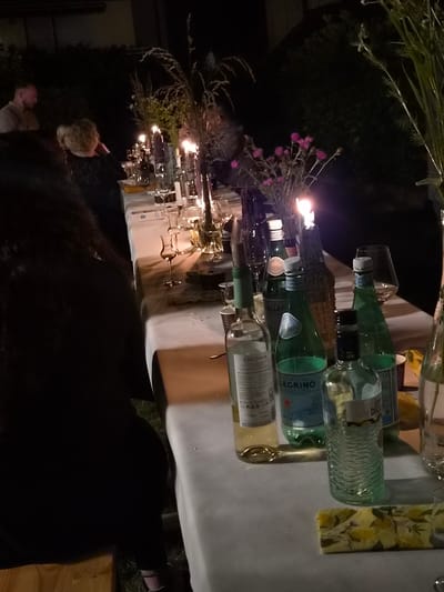 The image depicts an outdoor evening gathering, likely a dinner or party, set in the city of Bettembourg, Luxembourg. A long table, draped with a white tablecloth, is laden with various bottles of beverages, including wine and sparkling water. Among the prominent brands visible are "San Pellegrino" and "Ciconia." Several wine glasses and other glassware are scattered across the table, suggesting active use. Candles are lit, casting a warm glow and contributing to the ambiance. In the background, a person with short, curly blonde hair is visible, facing away from the camera and appearing to be engaged in conversation or observing the scene. Another person, with short hair and wearing a light-colored shirt, is partially visible in the far left of the frame, also appearing to be seated. The setting is illuminated by artificial light and the flickering flames of the candles. The presence of decorative greenery and flowers, possibly wildflowers, adds a natural element to the arrangement. The overall impression is one of a relaxed social event taking place in a garden or patio area.