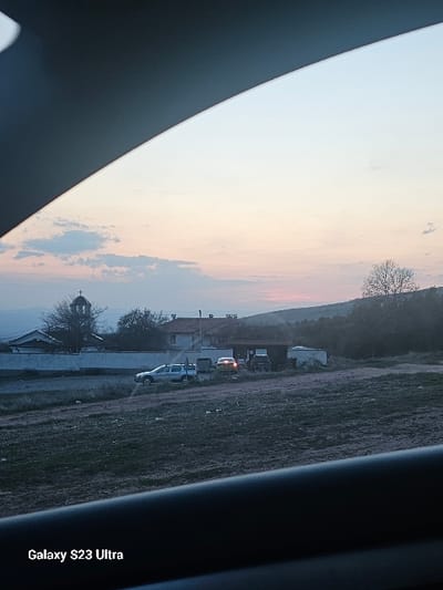 The image depicts an outdoor scene in Balsha, Bulgaria, captured from inside a vehicle during either sunrise or sunset, indicated by the sky's light blue, pink, and orange hues with scattered clouds. The foreground consists of a dark, uneven, sparsely vegetated field. In the midground, a light-colored SUV or pickup truck is parked, facing left. Beyond this, a series of structures includes a church with a distinct bell tower and a visible cross on its apex, situated next to a white-walled building with a terracotta roof. A long, light-colored wall or fence separates the field from these buildings. To the right of the church, two additional vehicles are parked near a smaller, dark-roofed structure; one is a yellow or orange vehicle with illuminated headlights facing right, and a darker vehicle is partially visible alongside it. The background features silhouetted trees and hills against the horizon. No individuals are visible. The watermark "Galaxy S23 Ultra" is present in the lower left corner.
