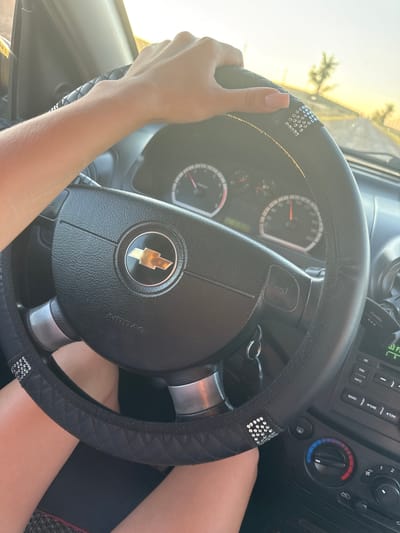 A close-up, eye-level shot shows a person's right hand resting on the steering wheel of a car. The steering wheel is black with a diamond-stitched cover and adorned with small, clear rhinestones. The Chevrolet logo is visible in the center of the steering wheel. The car's dashboard instruments, including a speedometer, are visible in the background. The sun is setting, casting a warm glow and visible through the windshield, creating a slightly overexposed effect. The environment outside the car appears to be a rural or suburban area, with a tree and telephone pole visible in the distance. The image was captured in the city of Iordanovca, Moldova.