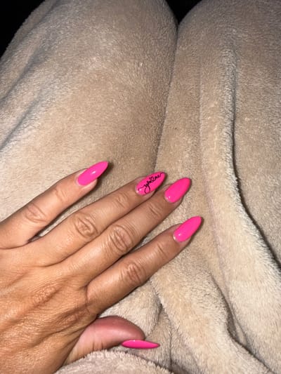 The image shows a person's hand with bright pink, almond-shaped acrylic nails resting on a soft, beige, fuzzy blanket. The hand is positioned in the lower right quadrant of the frame. The lighting suggests an indoor setting with soft, ambient light, possibly in the evening or a dimly lit room. The focus is primarily on the nails and the texture of the blanket.

Notable details include a black cursive script design on the ring finger nail, which appears to spell out "RubyLane". The skin on the hand shows some texture and is tanned. The blanket is folded, creating soft folds and shadows, contributing to the cozy and intimate feel of the image. No specific time of day or weather cues are discernible, and the location is given as Fairview Township, United States of America. The overall impression is one of personal care and attention to detail, with the vibrant nails standing out against the neutral background.