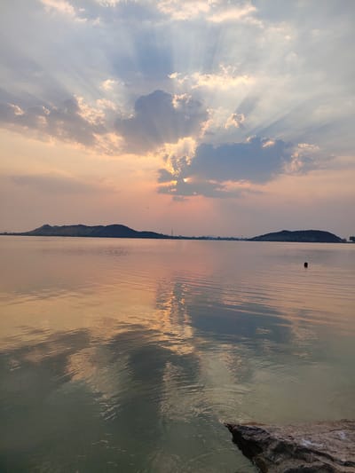 The image depicts a serene landscape at sunset in Kammavaripalli, India. A large body of water dominates the foreground, reflecting the colors of the sky. Two prominent hills are visible in the distance. The sky is filled with clouds, and rays of light emanate from behind them, creating a dramatic effect. A dark object, possibly a person's head, is visible in the water. A rock is visible in the bottom right corner of the image. The water is calm with minimal rippling.