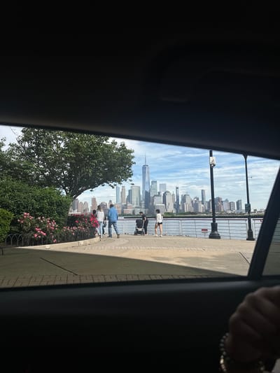 This image captures a view of the New York City skyline, particularly the World Trade Center, from Jersey City, New Jersey, United States. The photo is taken from inside a vehicle, as indicated by the framing of the car door and interior.
Outside the car window, pedestrians are visible on a waterfront promenade. There are flowers and vegetation along the railing of the promenade. The weather is sunny, with some clouds in the sky.
