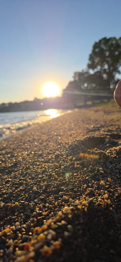 The image captures a close-up, low-angle view of a pebble-strewn beach during sunset. The sun is low in the sky, casting a warm, golden glow on the scene and creating bright reflections on the water. The pebbles in the foreground are sharply focused, showcasing their varied colors and textures, while the background, including the water, distant shoreline, and trees, is blurred due to the shallow depth of field. The sky is a clear, light blue transitioning to a soft yellow near the horizon where the sun is setting. The setting appears to be a natural shoreline, consistent with locations around Issyk-Kul Lake in Kyrgyzstan, known for its beaches and scenic sunsets. There are no visible people or specific man-made objects, suggesting a natural, tranquil environment.