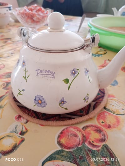 A white enameled teapot decorated with small blue flowers is placed on a patterned circular mat on a table covered with a yellow tablecloth featuring fruit illustrations. The teapot has a rounded body, a lid with a knob, and a spout. A brand name "Tulvano kitchenware" is visible on the side of the teapot. In the background, there is a glass bowl filled with a pink salad and other tableware. The scene appears to be an indoor setting, possibly a dining table. The bottom left corner of the image shows "POCO C65" and the bottom right corner shows "30.07.2026 12:39". This image is not geotagged to Beruniy, Uzbekistan.