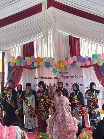A group of young people, predominantly female, are standing on a stage under a tent with white and maroon fabric draping. They are dressed in formal attire and many are wearing sashes and medals, suggesting an awards or graduation ceremony. A banner behind them reads "Akhirussanah, Pentas Seni & Tahfiz Al-Qur'an" which translates to "End of Year, Art Performance & Qur'anic Memorization". The event appears to be taking place in Sukabakti, Indonesia, as indicated by the text on the banner and a logo that includes "SOB AZ-ZAHRA" and "CIBUG-TANGERANG". Some individuals are holding microphones and looking towards an audience. Balloons are clustered above the banner.
