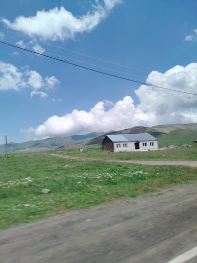 In the city of Jamshlu, Armenia, a long, low-profile building with a light gray metal roof and white brick walls stands amidst a vast, green, grassy field. The building has a dark entrance with a small overhang and several evenly spaced windows. To the left of the building, a dirt path winds through the field, and a tall, dark utility pole is visible near the path. In the distance, rolling green hills form a backdrop under a bright blue sky dotted with fluffy white clouds. Power lines stretch across the sky above the scene. The foreground shows a blurred section of a gray, paved surface, suggesting the image was taken from a moving vehicle.