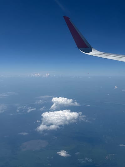 An aerial view captures the blue sky and clouds from an airplane window, with a portion of the aircraft's wing visible in the upper right quadrant. The wing, white with a red stripe on its tip, extends towards the center of the frame. Below the wing, a vast expanse of the earth's surface is visible, characterized by a mix of land and water bodies shrouded in haze and dotted with clouds. There are several distinct cloud formations, some appearing as fluffy white clusters and others as a thin, hazy layer. The land below displays varying shades of green and brown, suggesting a landscape with vegetation, possibly forests or fields, and hints of rivers or lakes. The image does not contain any information that can be used to determine a specific location within Mnogoozernyi, Russia, or any other city.
