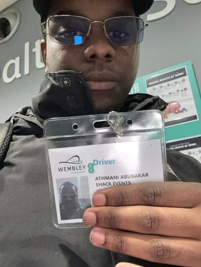 The image, rotated 90 degrees clockwise, presents a close-up portrait of a person, likely a man, with dark skin and dark hair, wearing rectangular, reflective sunglasses and a dark jacket. He is holding up an identification card in a clear plastic holder, displaying it towards the viewer with a neutral expression.
The ID card clearly identifies the individual as "ATHMANI ABUBAKAR" and indicates an affiliation with "SHACK EVENTS." It prominently displays the "WEMBLEY" logo and text that reads "Driver," along with "CONNECTED BY E" (likely referring to the EE mobile network). A small ID photo, consistent with the person in the main image, is embedded on the card. The person's fingers, with neatly trimmed nails, are visible as he holds the card.
In the background, a light blue-green poster is partially visible, containing text such as "MENTAL HEALTH AT WORK" and "LOOKING AFTER YOUR AL HEALTH AT WORK." Another partial word, "alt," is visible from what appears to be a separate sign. The indoor setting, bright lighting, and the "Wembley" detail on the ID card suggest the location is an event venue or workplace in Wembley, United Kingdom.