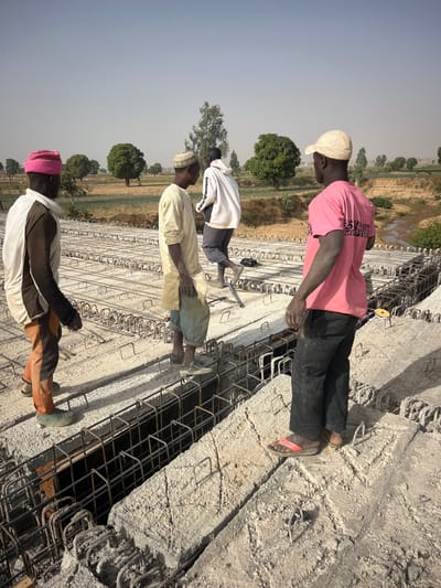 This image captures an outdoor construction scene under a hazy, light-blue sky, likely during the daytime. Four men, appearing to be construction workers, are visible on what seems to be a large concrete foundation or a bridge structure in various stages of construction. The ground is largely covered with an intricate network of steel rebar (reinforcing bars), suggesting preparation for concrete pouring.
From left to right (foreground to background, assuming the image is rotated upright):
1. In the foreground on the left, a man wears a pink knit beanie, a two-toned top (white upper section, dark lower section), and brown/orange pants with light-colored, possibly green, boots or shoes. He stands facing right, looking towards the construction work.
2. Behind him and slightly to the right, another man is dressed in a light-yellow or beige long garment, possibly a traditional tunic, and a patterned dark cap (kufi style). He wears light-colored shoes and also faces right, observing the scene.
3. Further back and to the right, a third man is seen wearing a white hoodie or jacket over dark shorts or pants, with dark shoes or boots. He is leaning forward or bending over, possibly engaged in a task involving the rebar structure on the ground.
4. In the background on the far right, a fourth man stands upright, wearing a pink short-sleeved shirt, dark pants, and a light-colored baseball cap. He has red and black sandals on his feet. He appears to be observing the work or taking a break.
The construction site features large sections of poured concrete mixed with exposed rebar, indicating ongoing work. The rebar forms grid-like patterns across the ground, hinting at the structural framework. Beyond the construction area, the background shows a landscape with dry, light-brown ground or fields, dotted with scattered green trees under the hazy sky. The overall impression is one of active construction in a warm, possibly rural, environment. No discernible text is visible in the image.