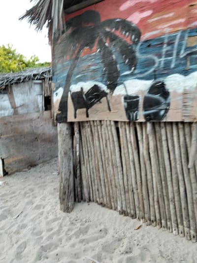 An exterior scene in Gombato Bongwe ward, Kenya, depicts two rustic structures on sandy terrain under a bright sky. In the immediate foreground to the right stands a shack with a weathered thatched roof. Its upper wall features a vivid mural painted on what appears to be wooden paneling, illustrating a sunset or sunrise with red, orange, and blue hues. A prominent black silhouette of a palm tree dominates the left side of the mural, with additional black silhouettes of beach furniture (possibly a lounge chair) and two beverage containers depicted on a simulated sandy surface below. Faint blue text or symbols are visible within the mural's blue section. The lower portion of this shack's wall is constructed from vertically arranged, light-colored wooden poles or thin logs, forming a screen, secured by a thicker, darker wooden post on the left. In the background to the left, a second, less distinct shack with a thatched roof is visible, appearing to be constructed from earth-toned materials, possibly adobe or mud, with a dark rectangular opening. The ground throughout the visible area is composed of light beige sand with minor undulations. Green foliage is visible in the upper left background. No individuals are present.