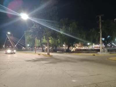 The image shows a street scene at night in Villa Carlos Paz, Argentina. A car with headlights on is driving on the left side of the frame. The road surface is asphalt with visible cracks and debris. The area is illuminated by streetlights, with one streetlight prominently displaying lens flare. Trees with hanging foliage line the street. In the background, a public area with benches and decorative lights is visible. A bridge structure with cable supports is present to the left. A utility pole stands on the right. The overall scene is a quiet urban landscape.