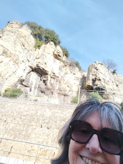 A smiling woman in sunglasses is framed against a background featuring a towering rock formation with sparse vegetation. The sky is clear and blue. A lower wall runs horizontally across the midground, separating the viewer from the rock face. Utility lines are visible along the wall. The woman's face fills the foreground, her gray hair framing her face.