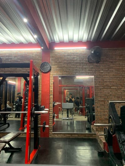 The image shows the interior of a gym in Kecamatan Kartasura, Indonesia, located at the coordinates -7.567 latitude and 110.757 longitude. The gym is equipped with weightlifting machines and other fitness equipment. There is a large mirror on the wall, reflecting the gym's interior and showing two people working out. One person is using a weightlifting machine and the other is standing in the background. The walls are made of brick, and the ceiling is made of metal panels. The floor is black and appears to be rubber. The gym is relatively empty, with only a few pieces of equipment in use. There are no other people in the image, but there are shadows of other people in the mirror. The room is well lit with fluorescent lights on the ceiling. The gym appears to be clean and well-maintained.