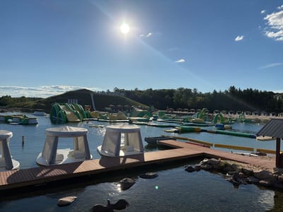 The image depicts an outdoor water park, likely located in Wisconsin Dells, Wisconsin, USA. The scene is set on a sunny day with a clear blue sky. The foreground features a wooden boardwalk extending into a clear body of water, with several white inflatable floating lounges with canopies docked alongside. In the water, there are various inflatable water structures, including slides, climbing walls, and obstacle courses, primarily in green and yellow. Some structures are partially submerged. The background shows a grassy hillside with a large sign that reads "CHULA VISTA NATURA." A few trees and structures, possibly part of a resort, are visible on the distant shoreline. The water appears calm, with reflections of the sky and structures.