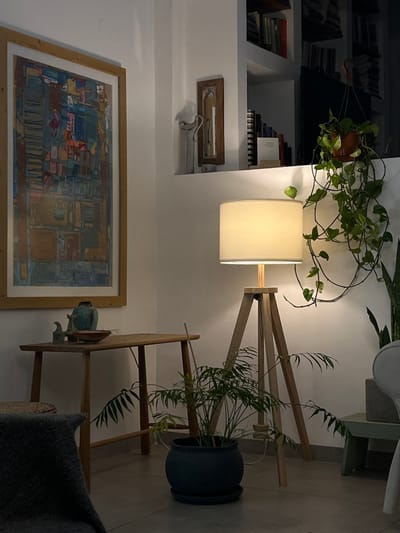The image depicts a living room in Alon HaGalil, Israel (latitude: 32.755, longitude: 35.219). The room features a wooden table with a vase and two small figurines, an abstract painting in a wooden frame, and a floor lamp with a white lampshade and a wooden tripod base. A potted plant is located on the floor next to the lamp and table. A vine plant, hanging from the ceiling, is present in the background,  alongside a bookshelf with various books. The room's walls are painted white. The image is taken from a seated perspective with a focus on the lamp, table, and plants. The scene is dimly lit with a soft glow coming from the lamp.