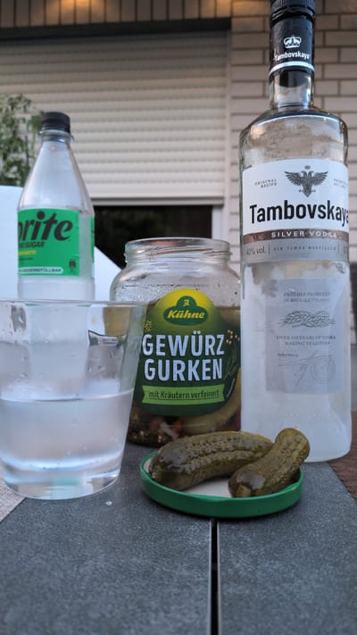 The image shows a close-up of a table setting outdoors, with a bottle of Sprite, a jar of pickled cucumbers, a glass of water, and a bottle of Tambovkskaya Silver Vodka. The background consists of a brick wall and a white rolling shutter. The scene appears to be set in Dissen am Teutoburger Wald, Germany, based on the presence of the Tambovkskaya vodka, a brand often associated with Russian traditions, and the Kühne brand of pickled cucumbers, which is a German brand. There are no people in the image, and the objects are arranged on a dark grey textured surface. The lighting suggests it is daytime.