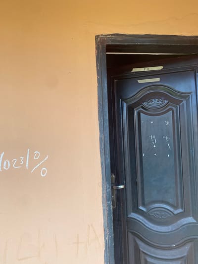 The image shows a closed, dark gray metal door on the right side of the frame. The door has an ornate design in the upper portion and a silver handle on the left side. Two pieces of yellow tape with writing are affixed to the door near the top. On the left side of the frame, on an orange wall, is writing in white chalk. The chalk writing appears to be a series of numbers and a percentage sign, "10231%". Faint chalk markings, possibly letters, are also visible lower down on the wall. The environment appears to be an outdoor wall in Bulamare, Nigeria.